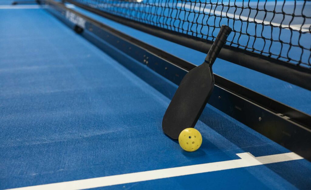 Pickelball racket and ball leaning against net on court